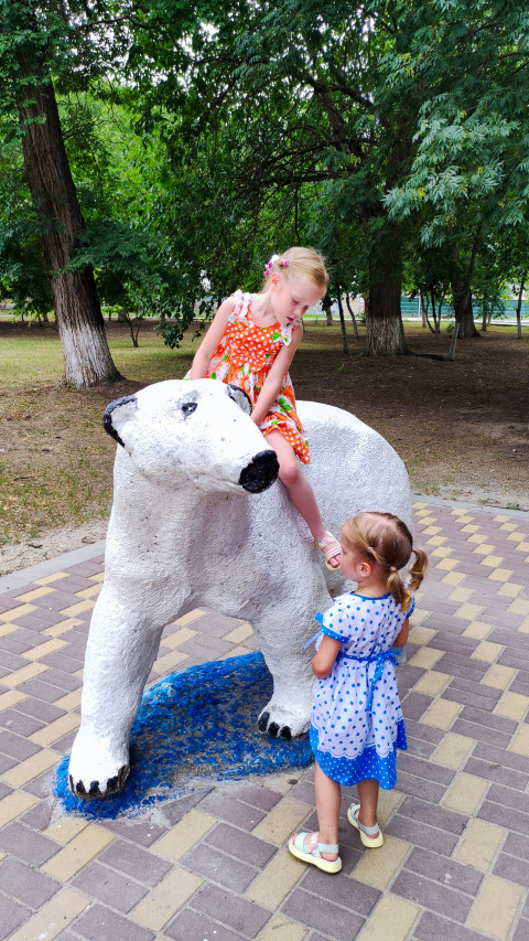 Две девочки играют у скульптуры белого медведя в парке