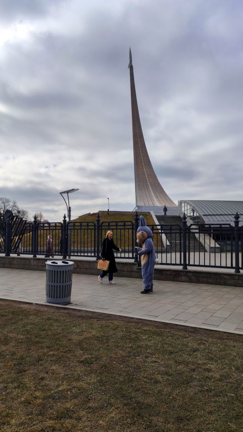 Люди идут по тротуару перед памятником Покорителям космоса на ВДНХ в Москве