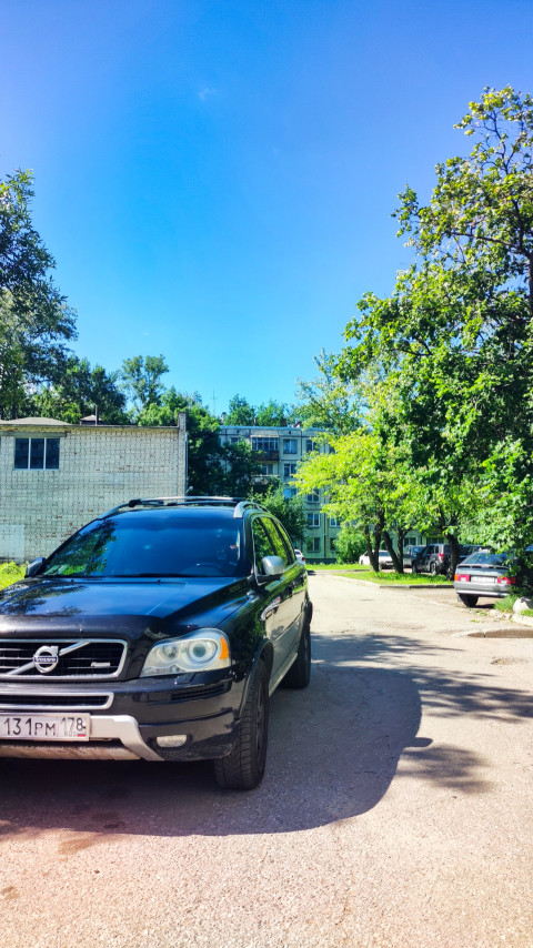 Чёрный Volvo SUV на дворовой дороге среди деревьев и жилых домов