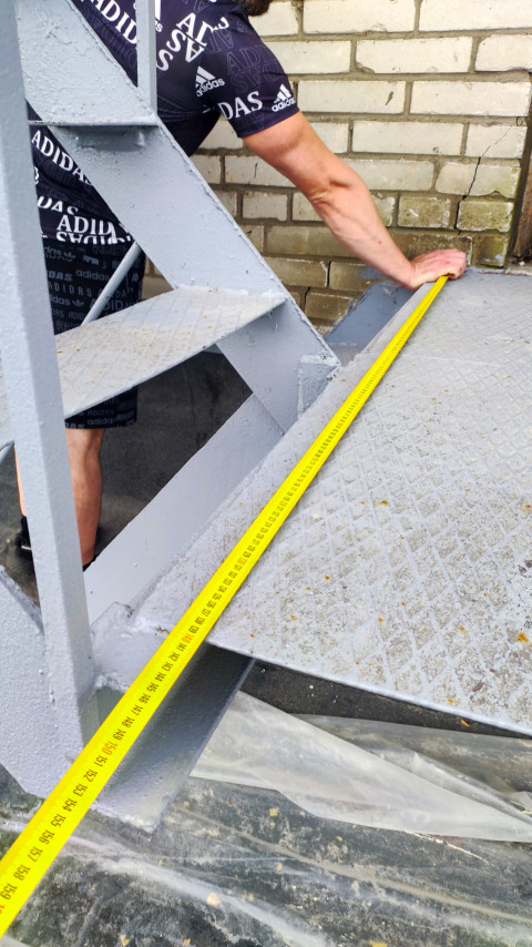 Рабочий измеряет рулеткой металлическую ступень у кирпичной стены