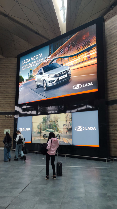 Пассажиры смотрят на гигантский экран с рекламой LADA в терминале