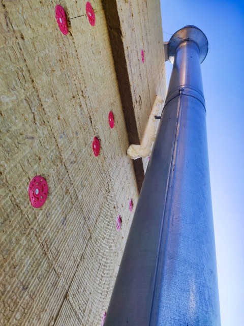 Водосточная труба рядом с утеплённой стеной, закреплённой тарельчатыми дюбелями, на фоне ясного неба